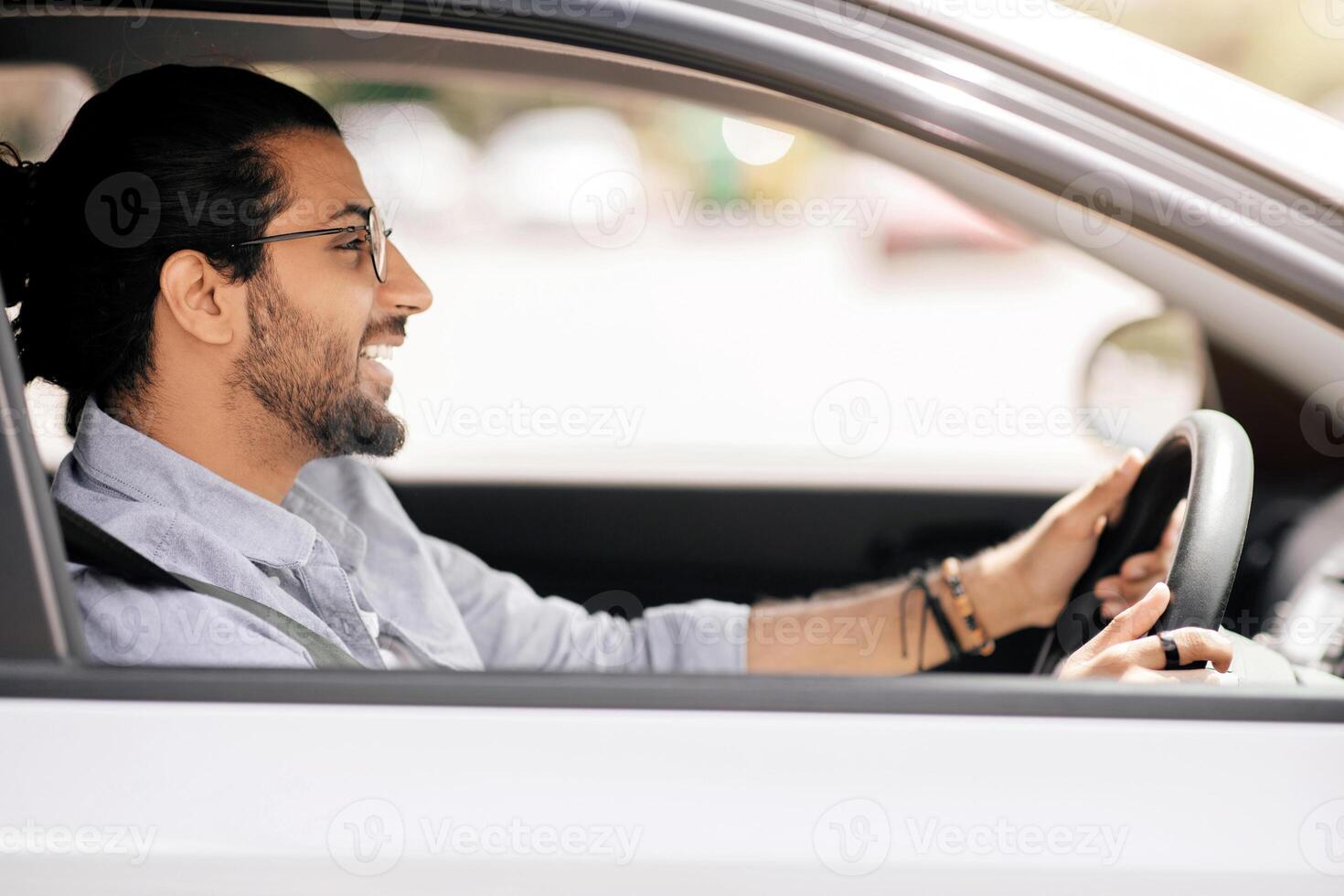 Handsome millennial long-haired arabic guy driving nice car, wearing glasses, side view, shot from outside, copy space. Cheerful indian man holding hands on wheel, looking at road and smiling photo