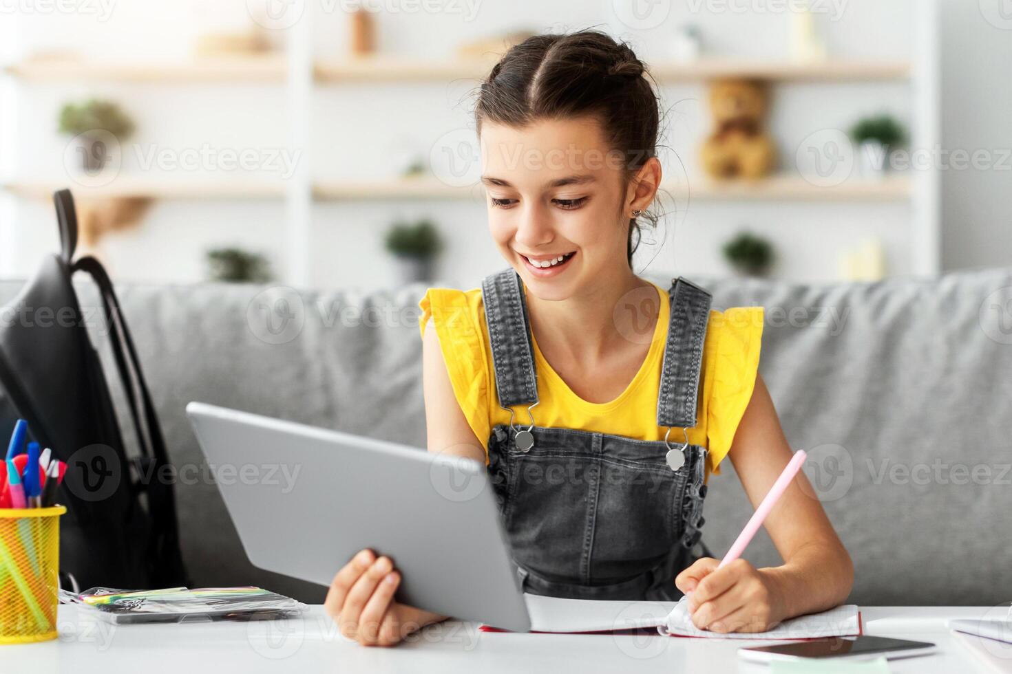 Children and Education Concept. Portrait of positive teen girl using doing homework writing in notebook sitting on couch at desk, holding and using tablet pad computer, enjoying distance homeschooling photo