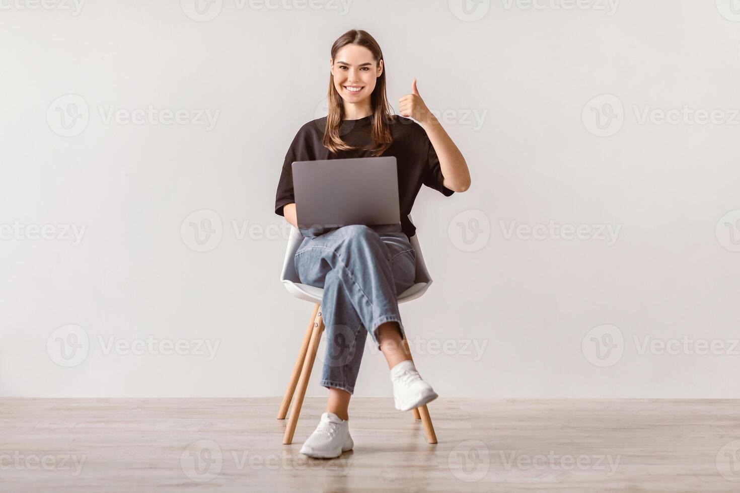 Smiling millennial woman working on laptop, showing thumb up gesture, recommending new website, sitting on chair against white wall. Young lady surfing internet, searching for information online photo