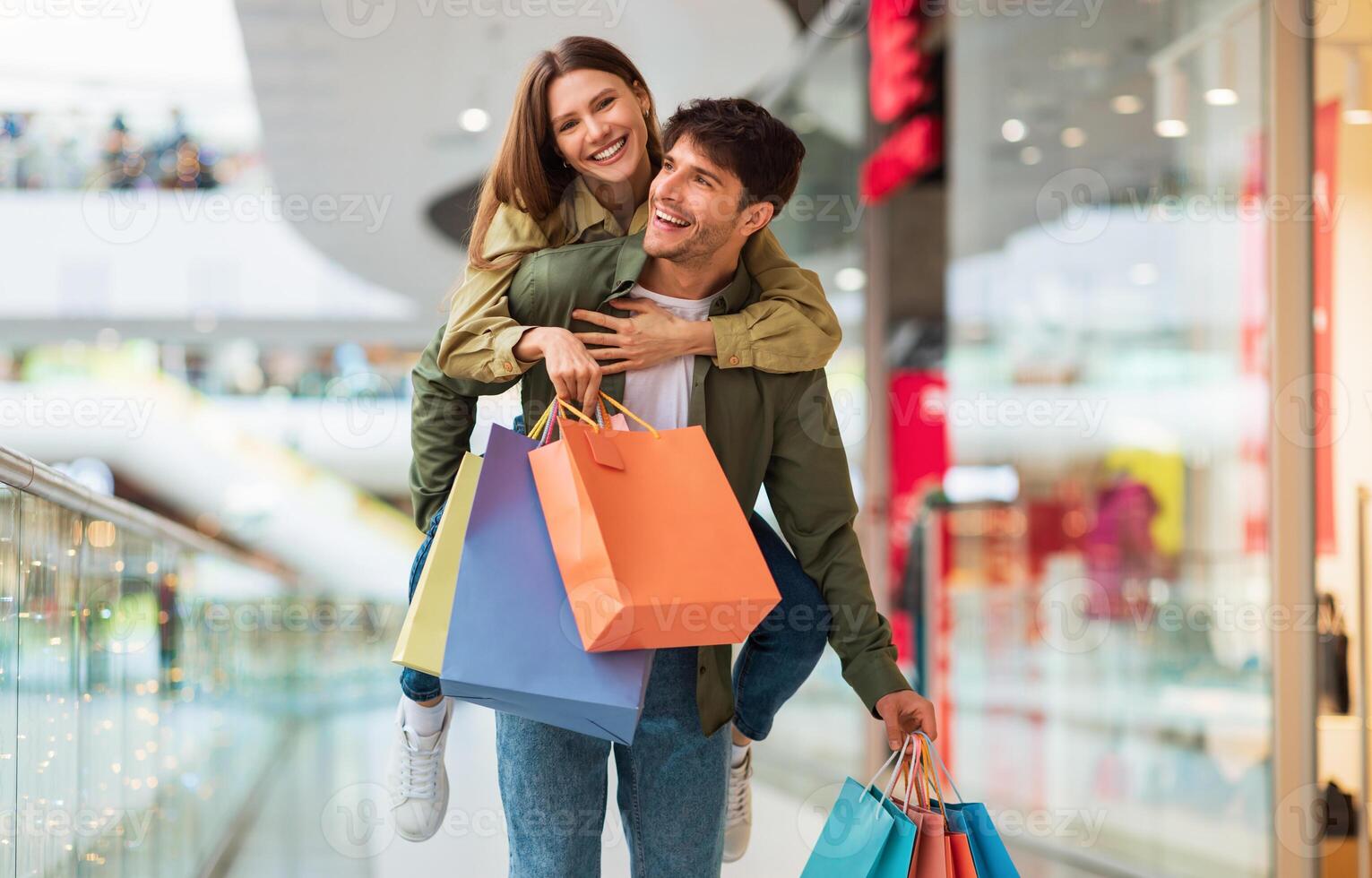Happy Spouses Shopping And Having Fun, Husband Carrying Wife Piggyback And Holding Colorful Shopper Bags In Modern Hypermarket On Weekend. Great Seasonal Sales And Commerce Concept photo
