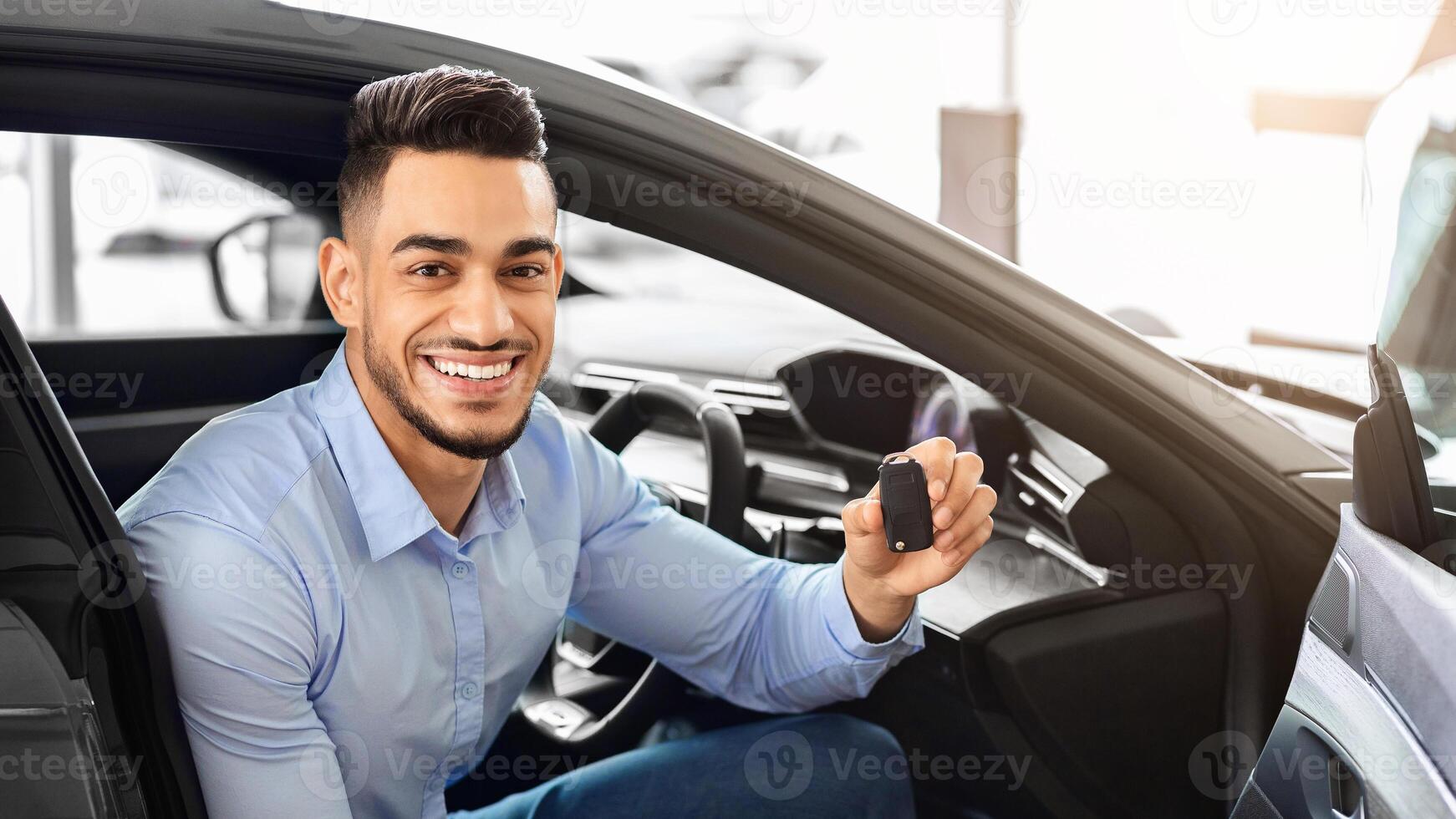 Handsome arab man customer sitting in auto and holding key, smiling at camera, buying car in showroom. Cheerful middle-eastern guy sitting inside nice sports car, happy to get new automobile photo