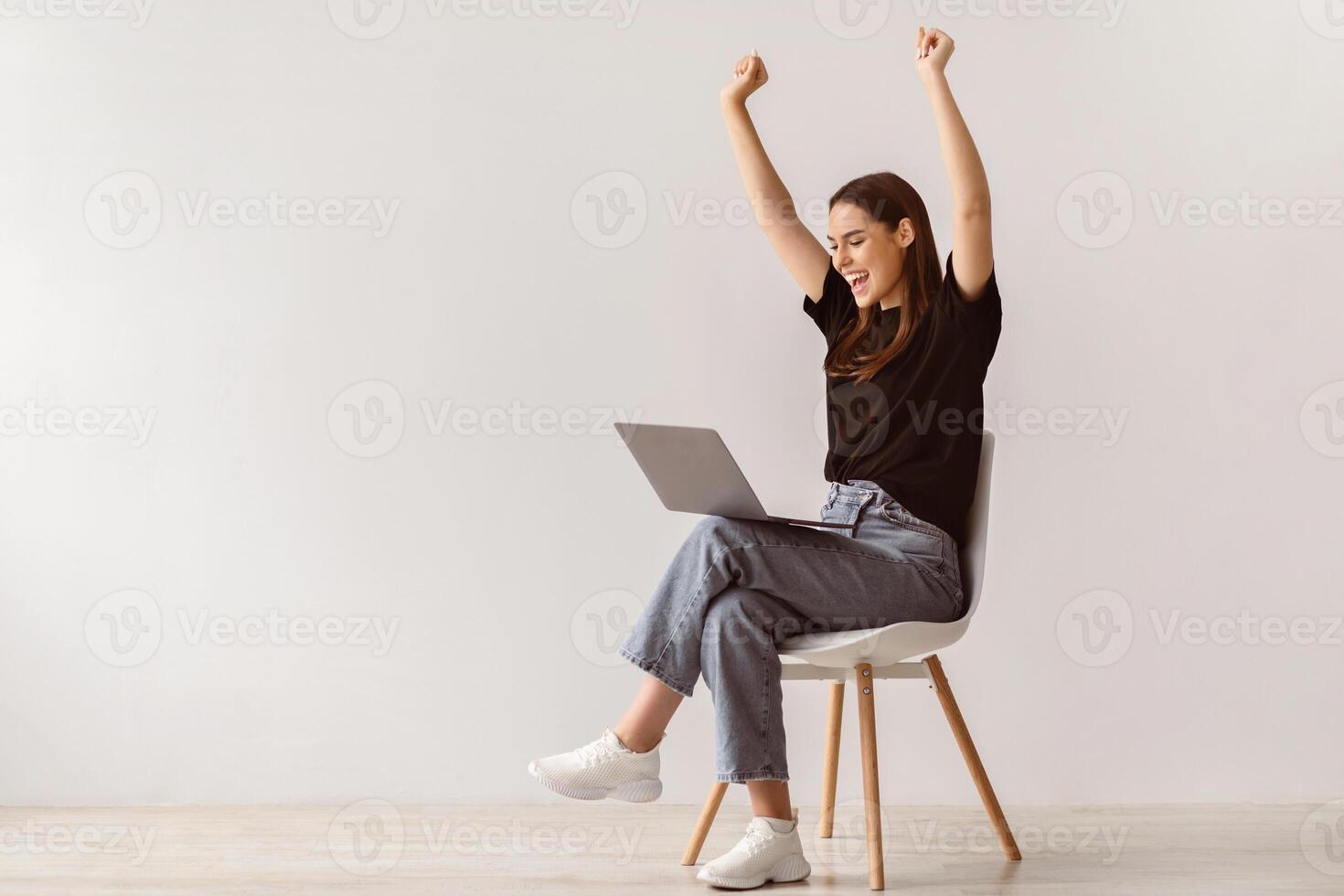 Entertainment and online. Happy young lady sitting on chair with laptop, raising her hands up, excited over huge lottery win or business success in studio, copy space photo