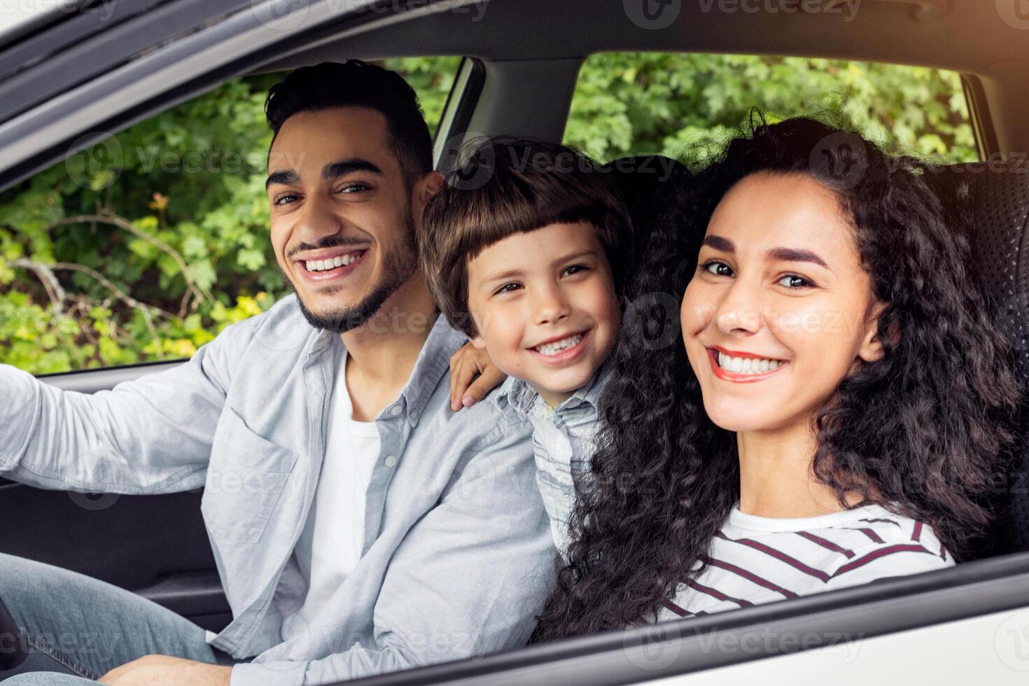 Cheerful glad millennial arab mother and father with kid go on vacation by auto and look at camera at open window. Family trip on new car, weekend together, love and relationship, positive emotions photo