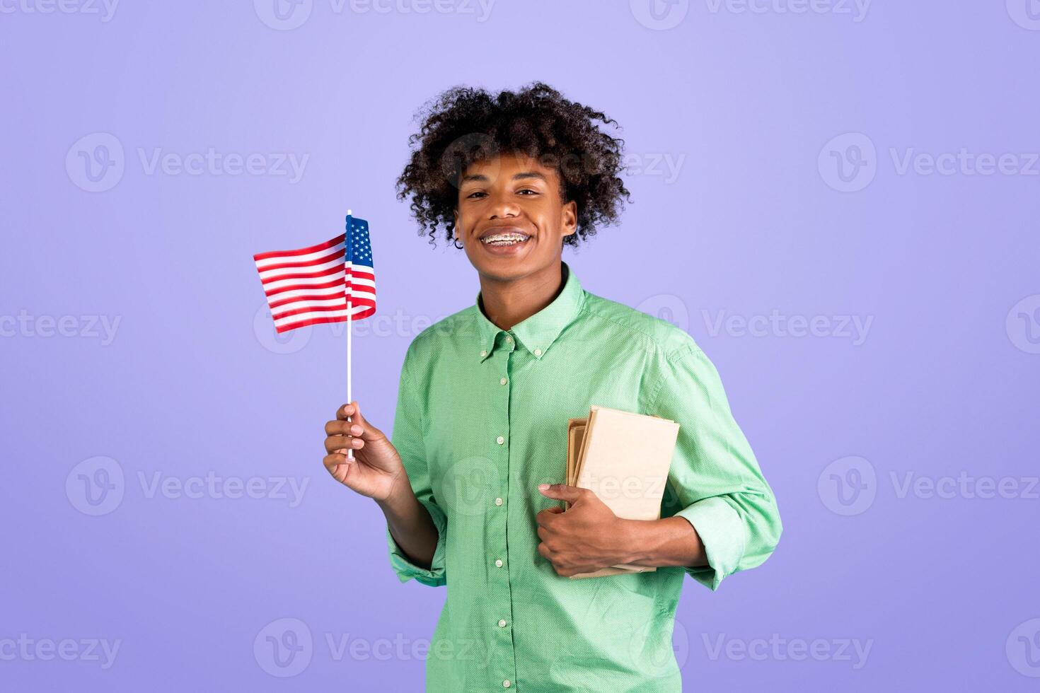 Student exchange and learning english. Zoomer happy black guy with books and small flag of USA go to american college, posing on blue background, studio shot, empty space photo