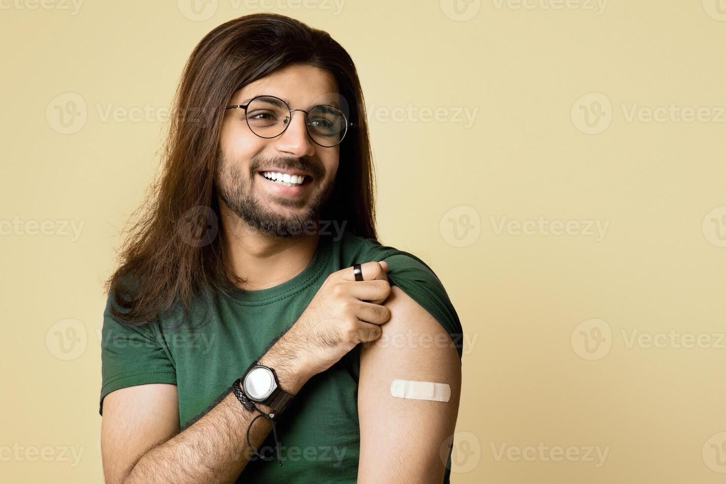 Happy millennial arab guy got vaccinated against COVID-19, showing shoulder with plaster on and smiling, sitting on chair over beige studio background, looking at copy space, panorama photo