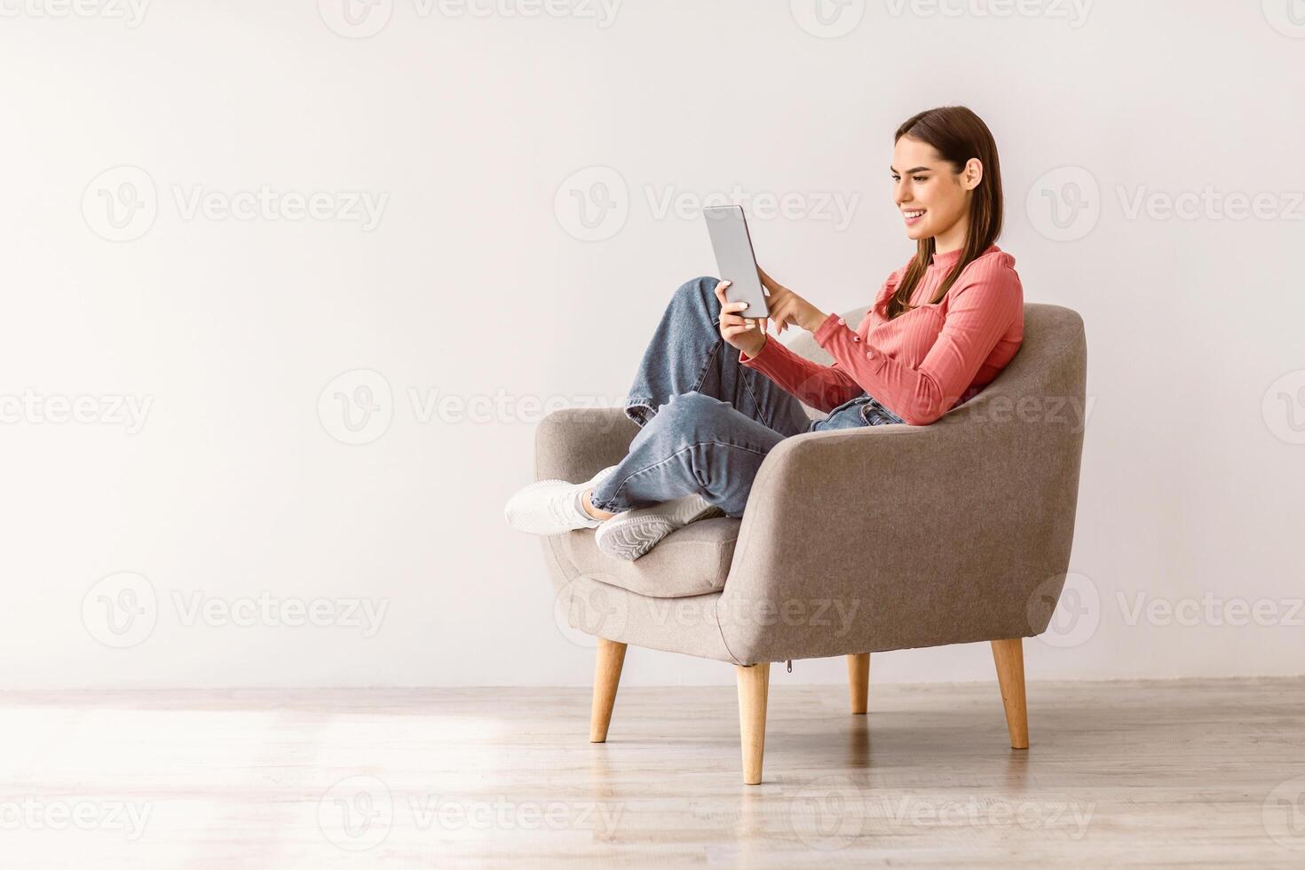 Happy millennial lady using tablet pc, studying or working remotely, having online conference, sitting in armchair against white studio wall, copy space. Young woman with touch pad checking new app photo