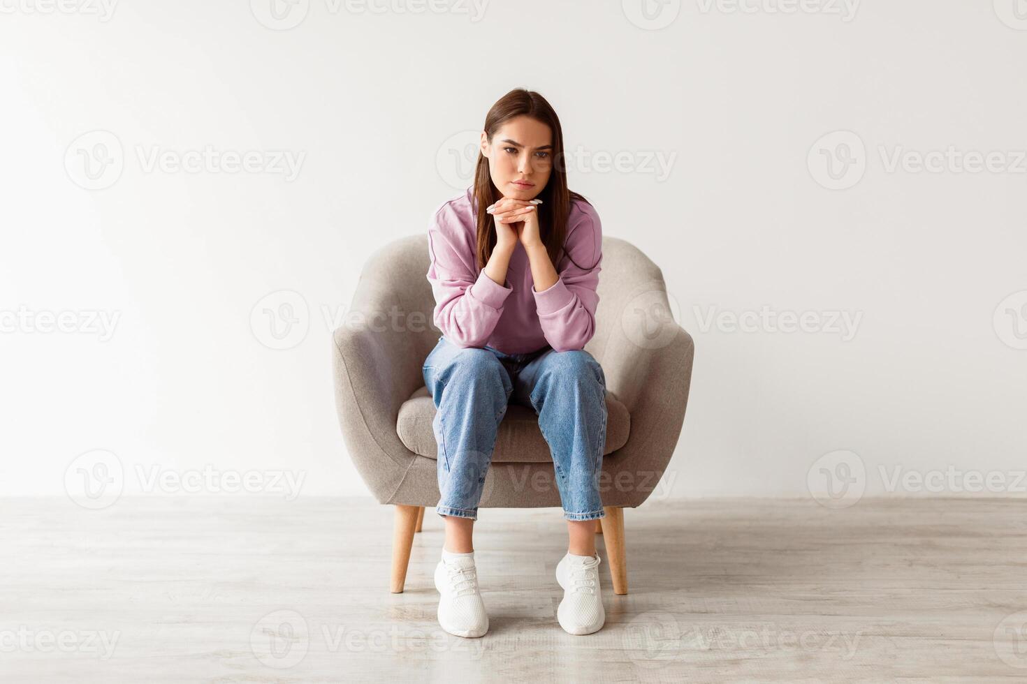 Negative emotions. Full length of young Caucasian woman sitting in armchair with dull or thoughtful face expression, feeling upset or tired, having problem or depression against white studio wall photo