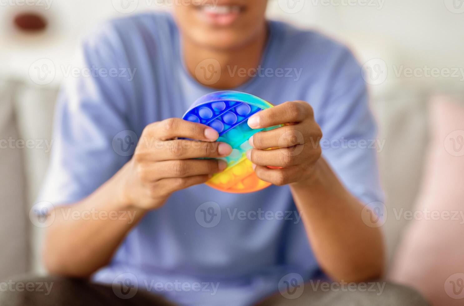 Closeup view of African American teenager playing with antistress POP IT toy at home. Unrecognizable black youth using sensory plaything to relieve stress or relax indoors photo