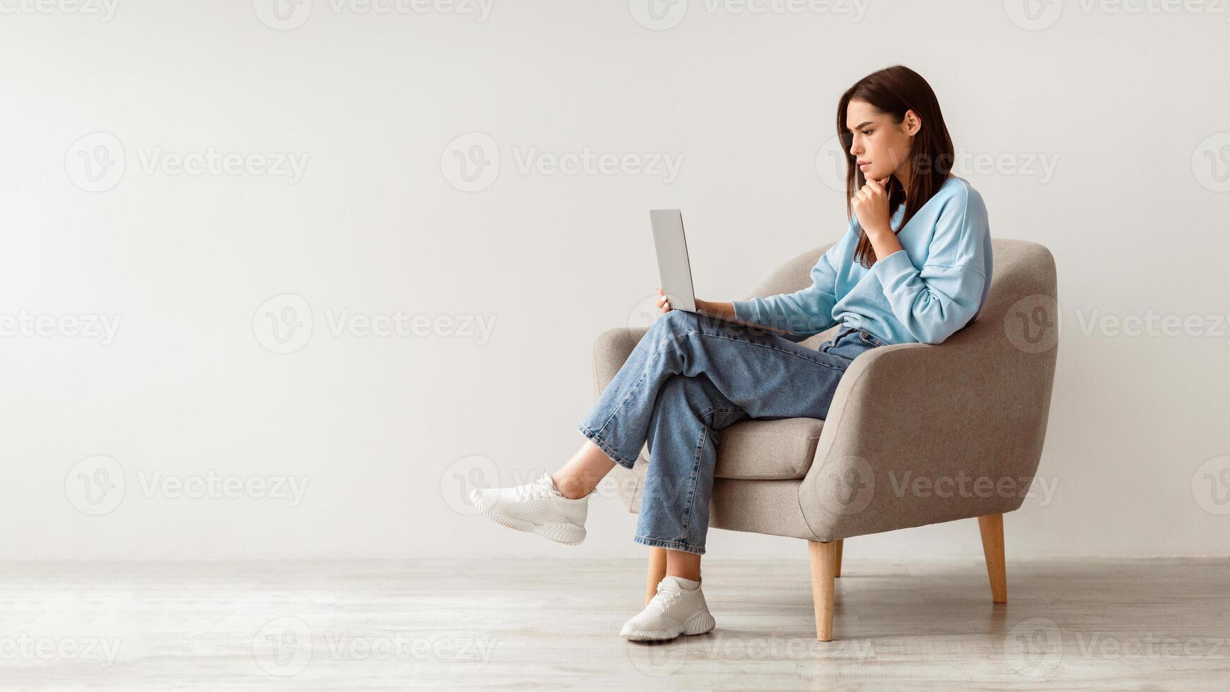Full length of serious young woman looking at laptop screen, sitting in armchair, having problem with online work or studies against white studio wall, banner with copy space photo