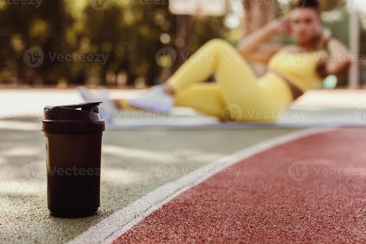 Black woman doing abs exercises crunches with arms behind head on floor yoga mat, working on six-pack abs in blurred background, selective focus on black shaker on basketball field ground photo
