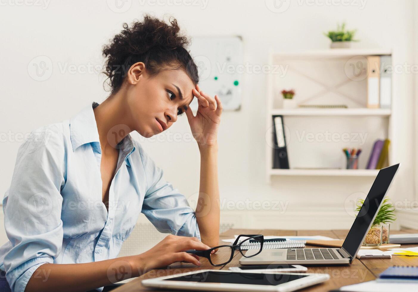 Deadline stress concept - sad african-american business woman sitting at desktop in office, working on laptop and holding hand on head. Hard working day, copy space photo