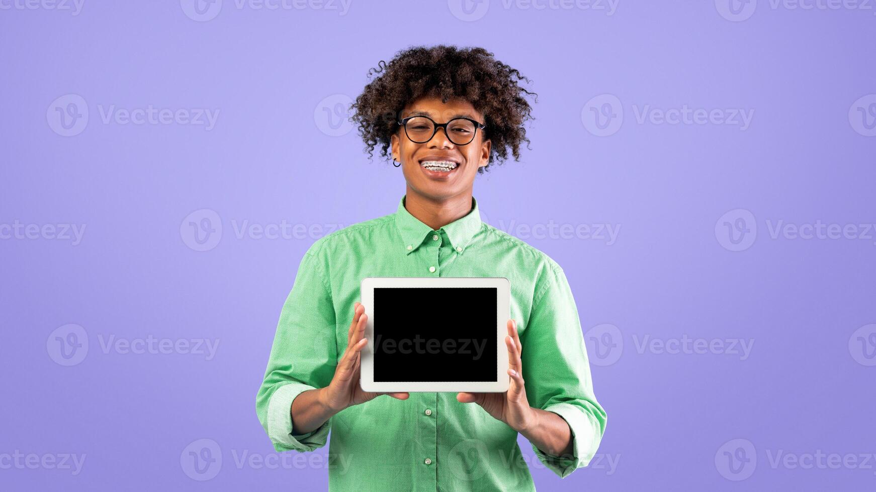 Happy african american teen guy showing new digital tablet with blank screen over blue studio background, happy teenager recommending newest mobile application for study online, copy space photo