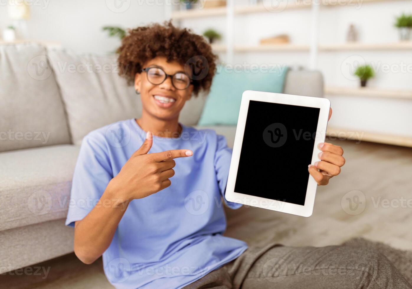 Cheerful black teenager pointing at tablet pc with blank screen at home, mockup for mobile app or website design. Online education, remote communication during coronavirus isolation photo