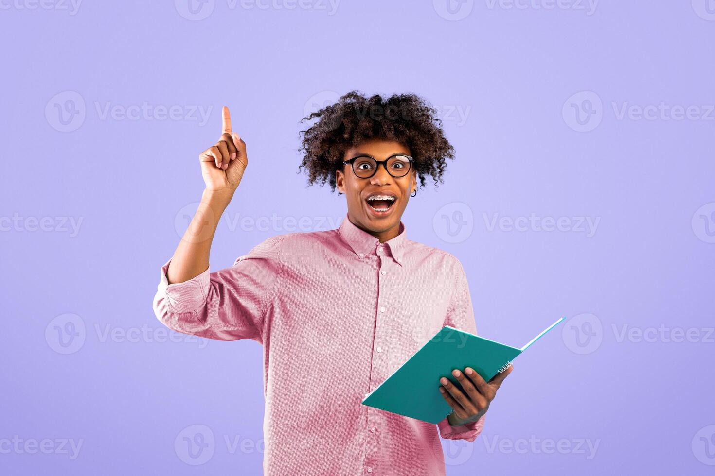 Happy african american male student pointing finger up, having creative idea, holding notebook on blue studio background. Black guy experiencing inspiration or eureka moment photo