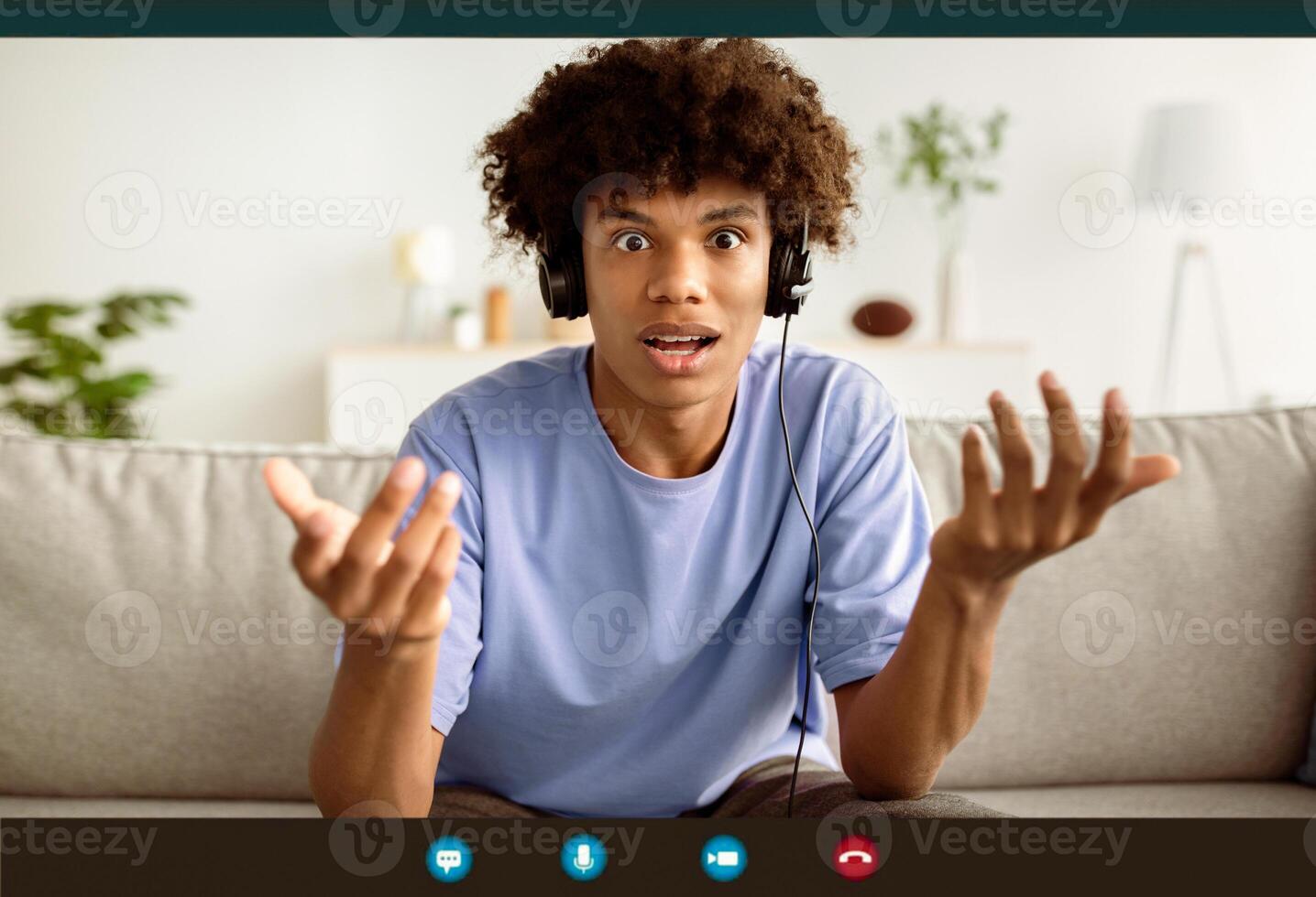 Webcam shot of shocked black teenager in headphones communicating online from home. Afro teen gesuring wtf, having call, feeling surprised during web conversation, computer screen view photo