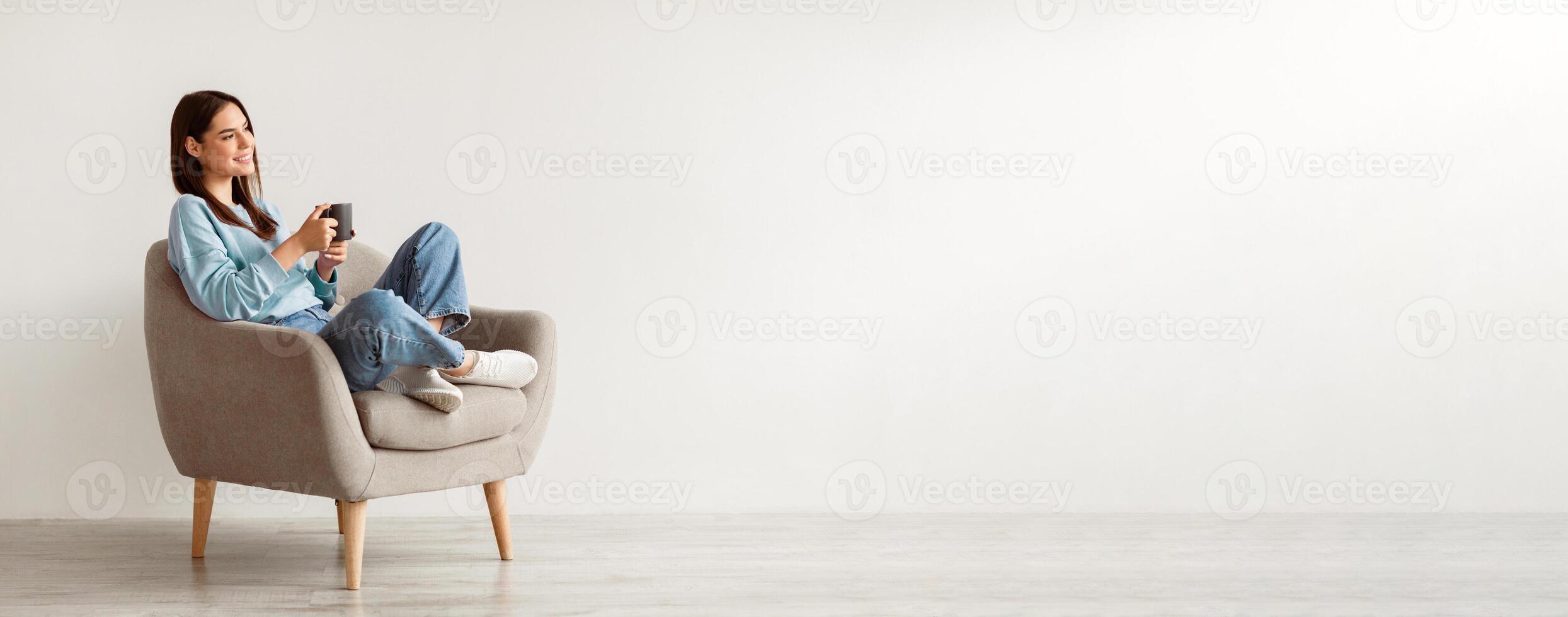 Cheerful young woman drinking aromatic coffee while chilling in comfy armchair against white wall, panorama with blank space. Smiling millennial lady having hot beverage, relaxing during her break photo