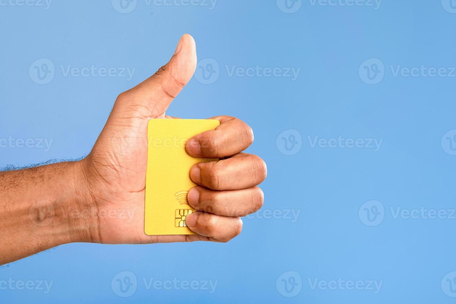 Cropped view of millennial African American man holding credit card and gesturing thumb up on blue studio background, empty space. Closeup of satisfied guy bank client recommending bank services photo