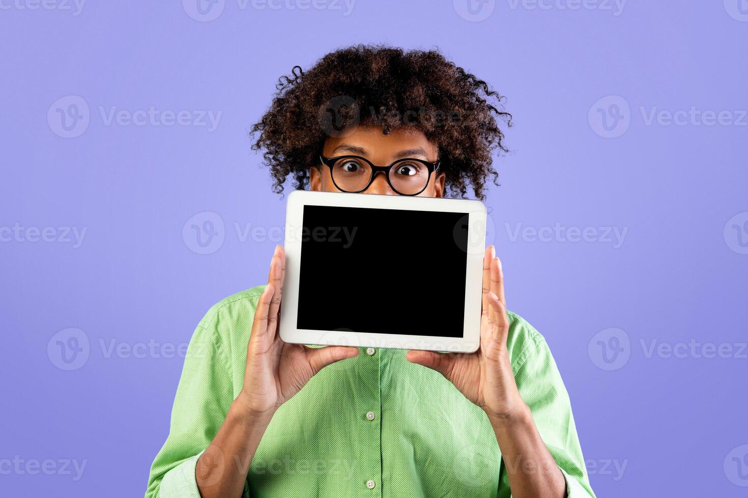 Amazed black guy in eyeglasses hiding behind digital tablet with empty screen over blue studio background. Excited teenager showing pad, mockup, advertisement for educational app photo