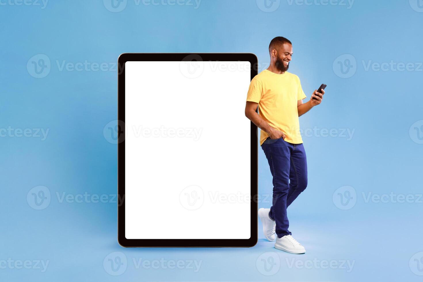 Optimistic young African American man leaning on huge tablet computer with blank white screen, using mobile phone on blue studio background, mockup for website or application design photo