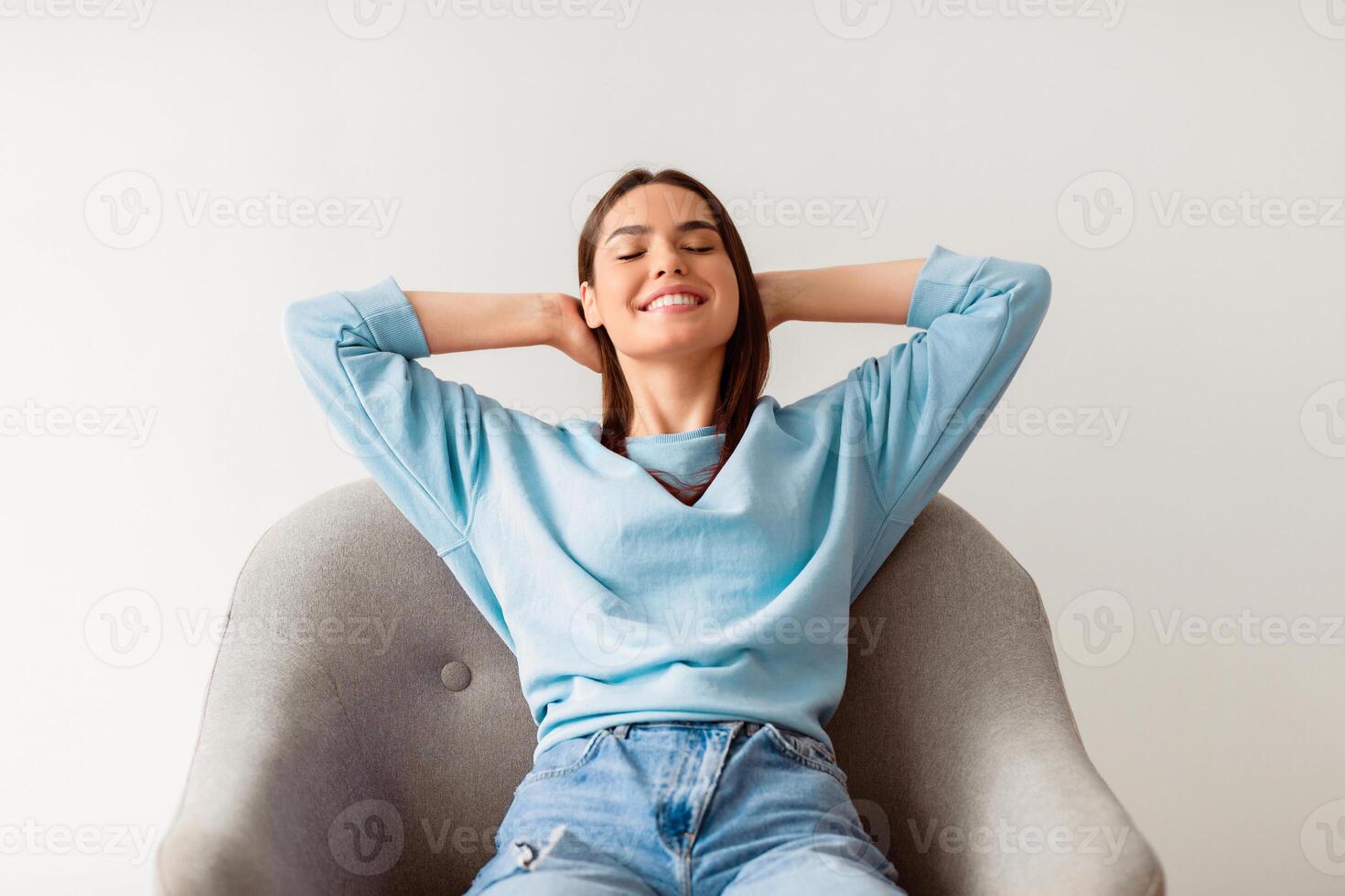 Pretty young Caucasian woman sitting in comfy armchair with hands behind her head, closing eyes, relaxing on white studio background. Millennial lady having rest, chilling, enjoying day off photo