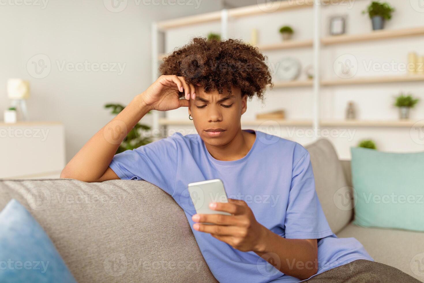 Funky black teen guy with mobile phone studying online, messaging with friend, posting in social network from home. African American youth talking to tutor on web, participating in remote meeting photo