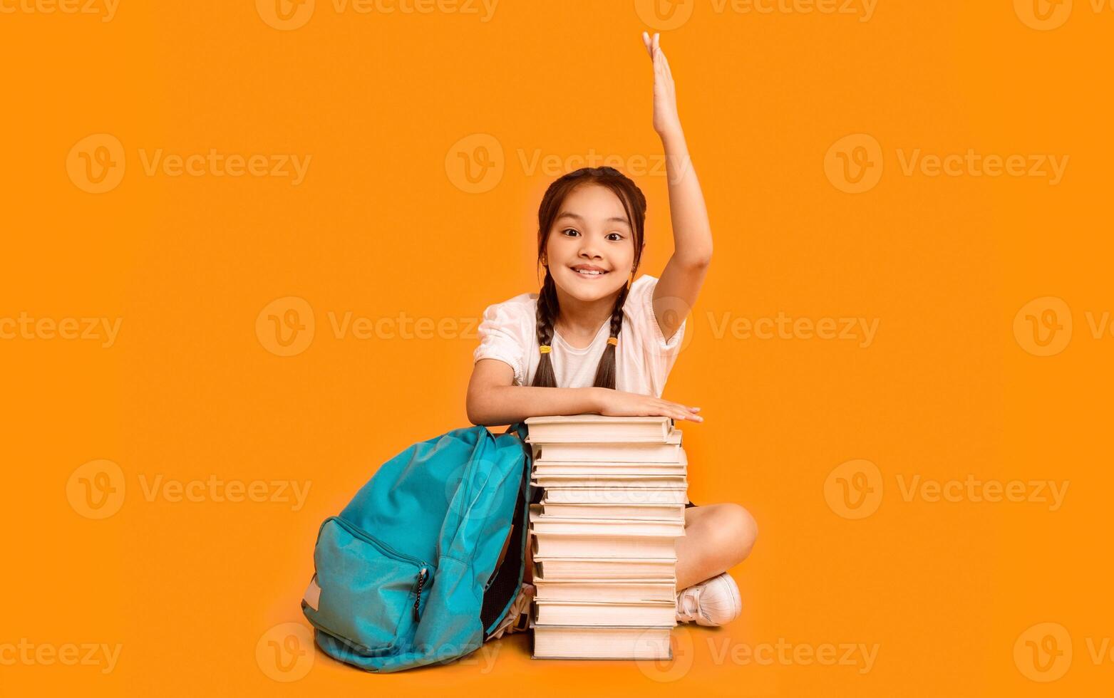 Clever Elementary School Girl Raising Hand Knowing Answer Sitting At Book Stack Over Yellow Background. Studio Shot, Empty Space photo