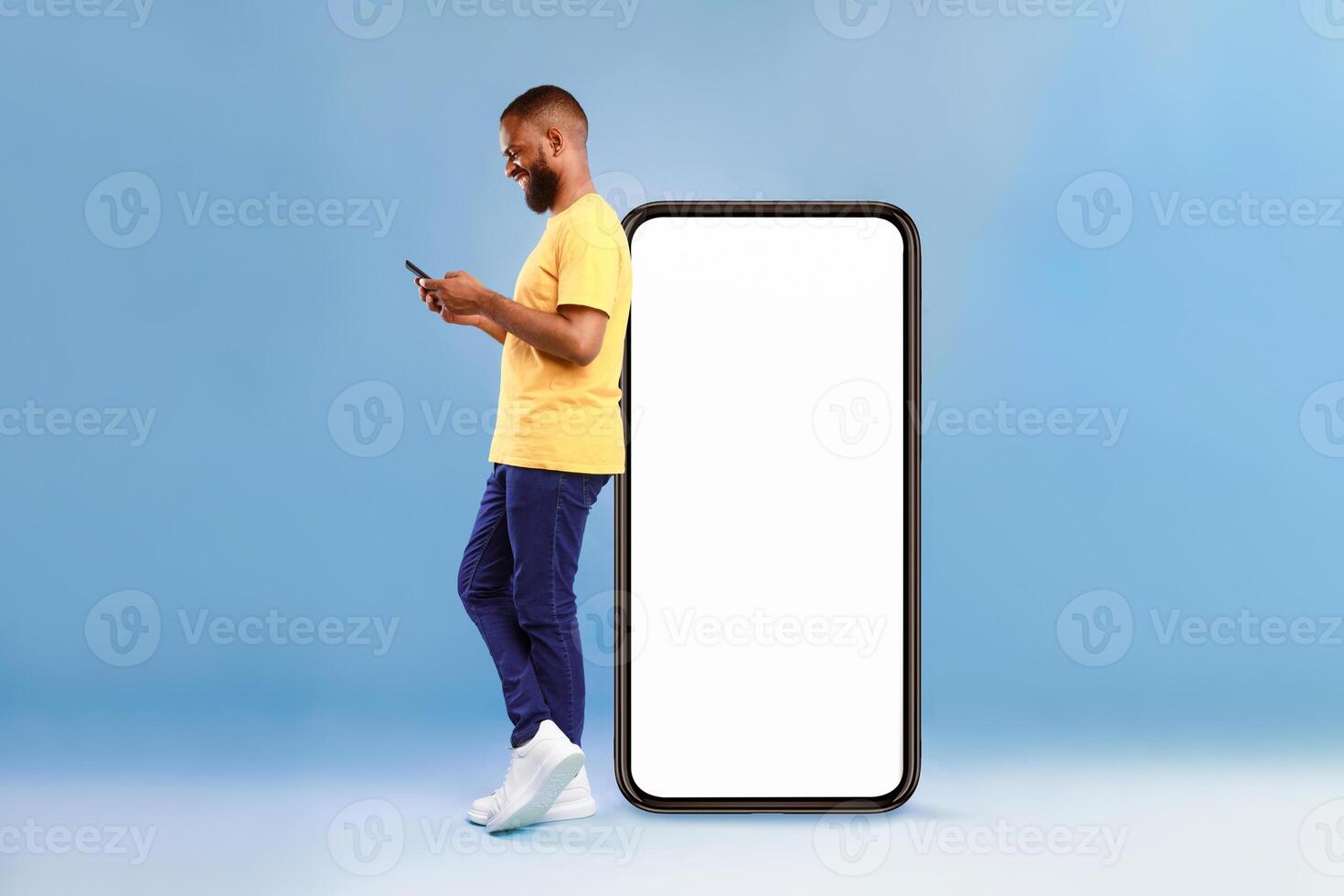 Full length of young African American guy using cellphone while leaning on giant mobile phone with empty white screen, blue studio background. Mockup for app or website, space for advertisement photo