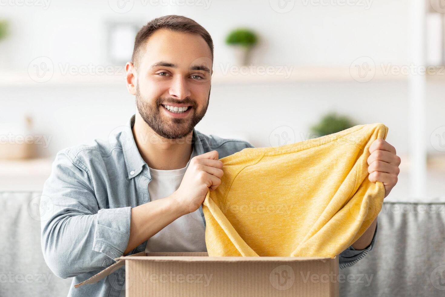 Happy Caucasian guy unboxing cardboard parcel, satisfied with successful shopping, taking out new sweater, receiving new purchase at home. Online store and delivery concept photo