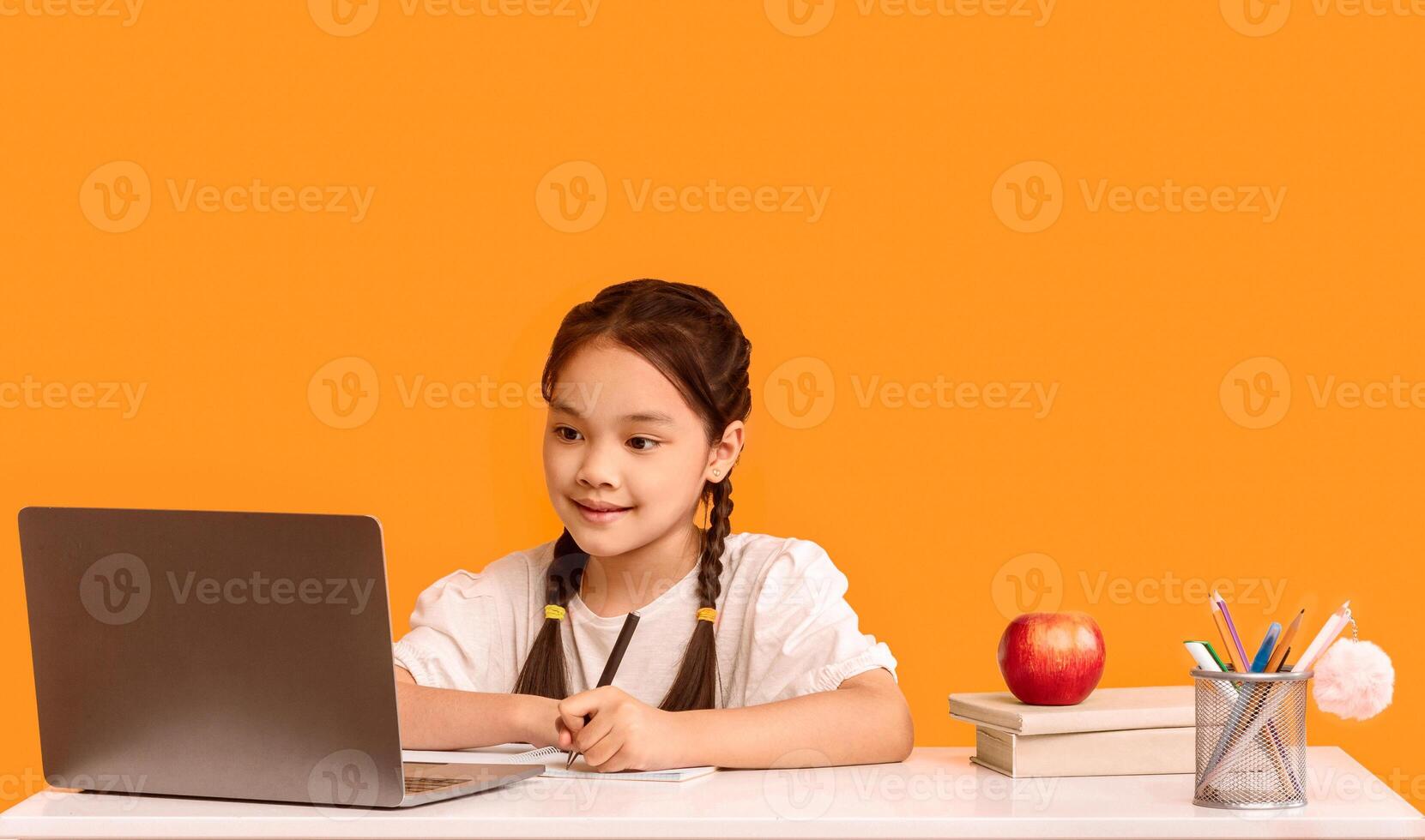 Adorable Japanese Elementary Student Girl Doing Homework At Laptop Computer Sitting On Yellow Background. Studio Shot, Free Space photo