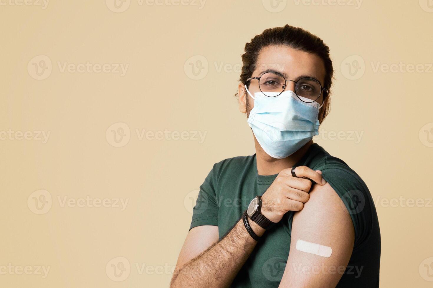 Long-haired arab man in protective face mask got vaccinated against COVID-19, showing shoulder with medical plaster on, sitting on chair over beige studio background, copy space, panorama photo