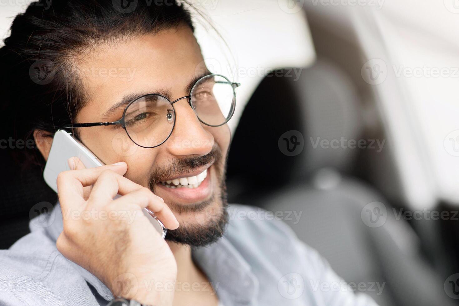 Closeup portrait of young cheerful indian man driver having phone call while driving car, side view, copy space. Happy middle-eastern guy enjoying summer trip, sitting in auto, talking on smartphone photo