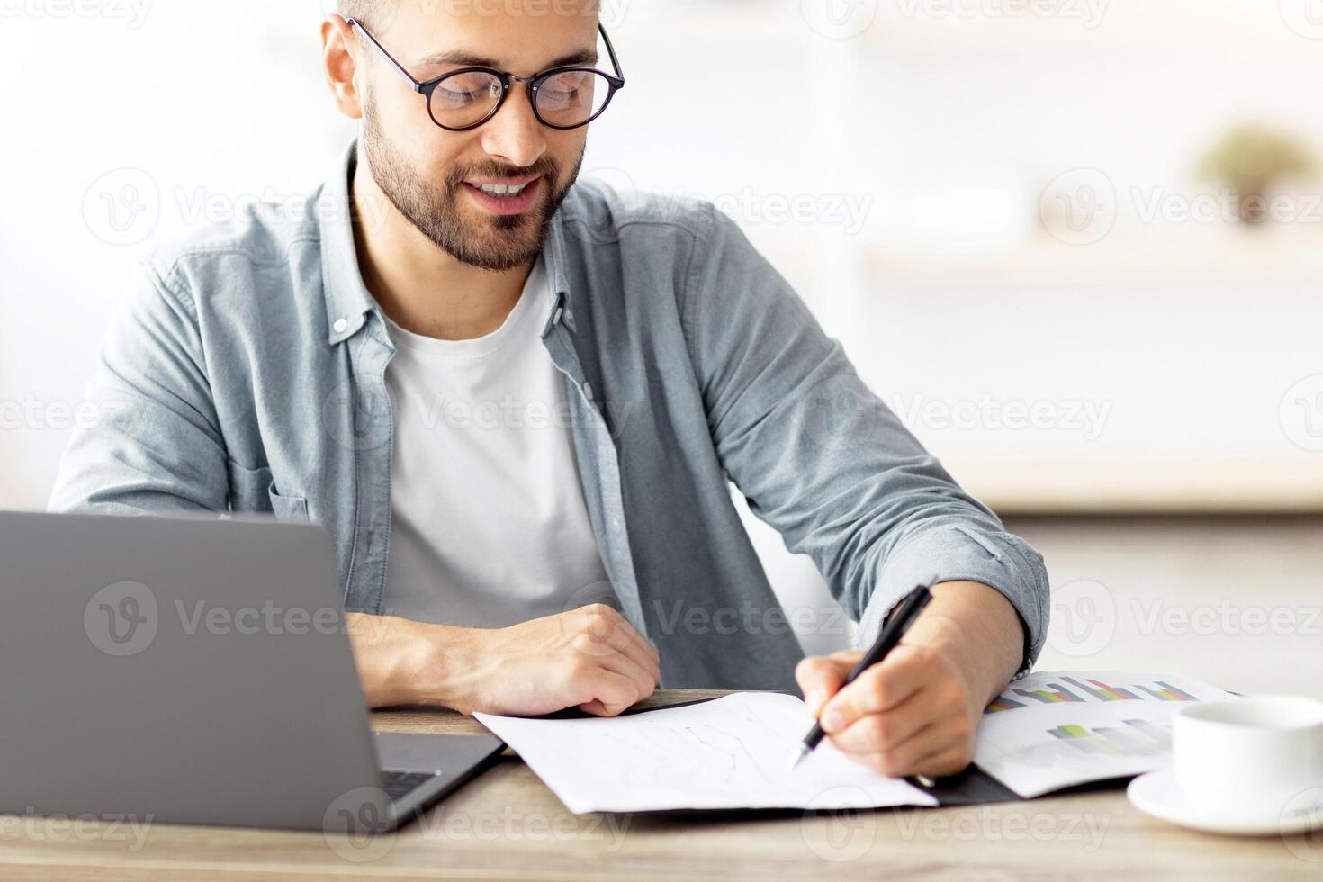 Young Caucasian businessman taking notes while working on laptop computer at home office. Freelancer noting down information from internet, planning working schedule, sitting at desk indoors photo
