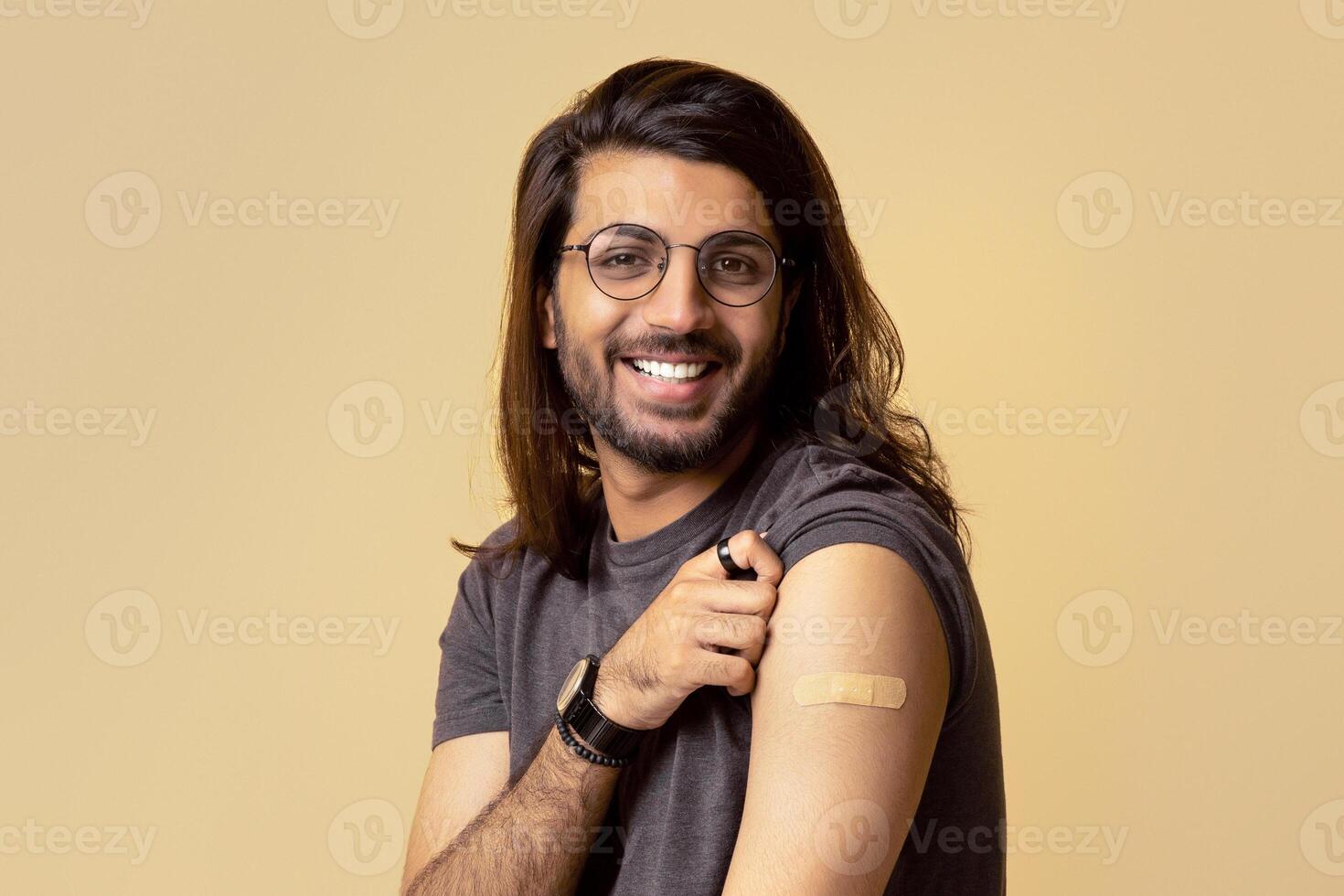 Happy long-haired arab guy in casual got vaccinated against coronavirus at hospital or clinic, sitting on chair over beige studio background, showing his shoulder with adhesive band on, copy space photo