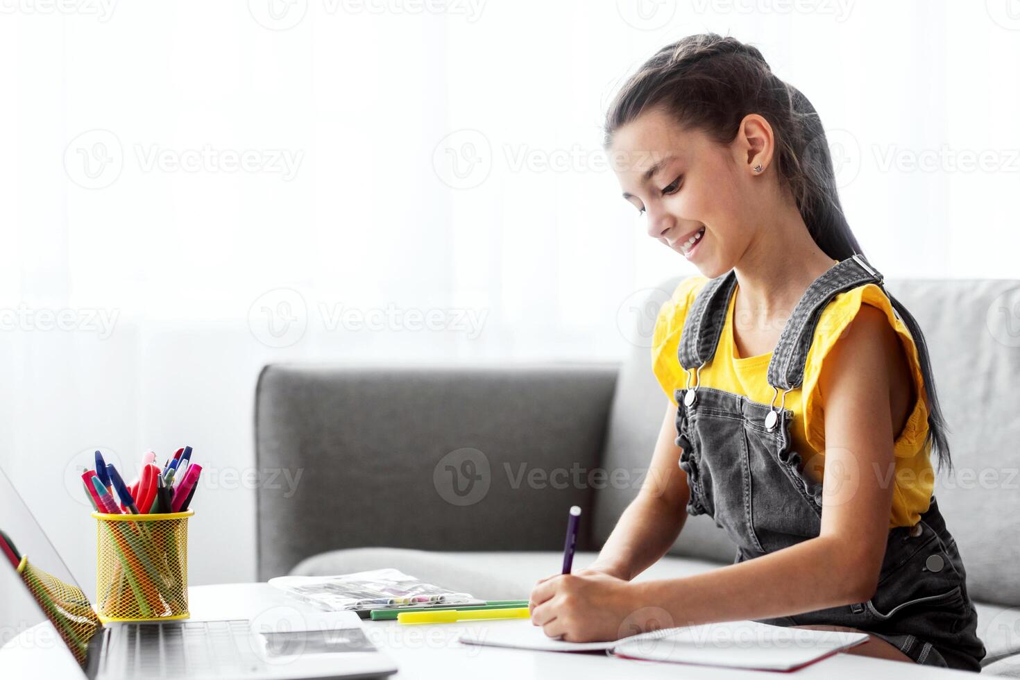 Portrait of smiling schoolgirl sitting on couch at desk with pc, writing essay or drawing picture in paper notebook. Pupil studying or taking notes in her diary, making checklist, free copy space photo