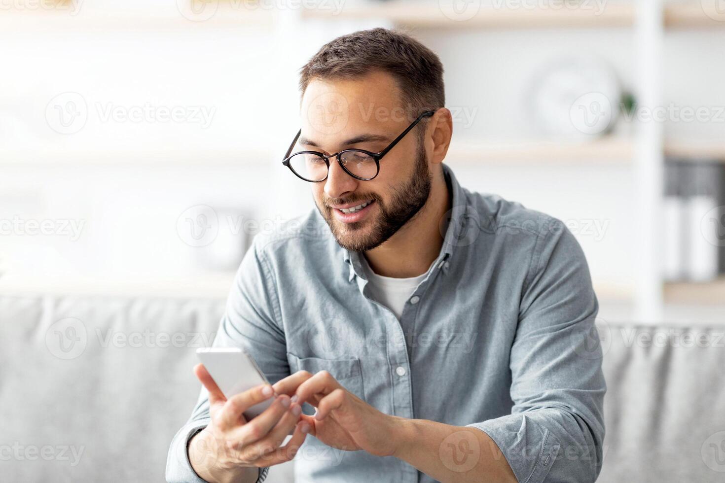 Positive young guy in eyeglasses sitting on sofa with mobile phone and texting to his friend, indoors. Millennial Caucasian man writing message, having call or broswing web at home photo