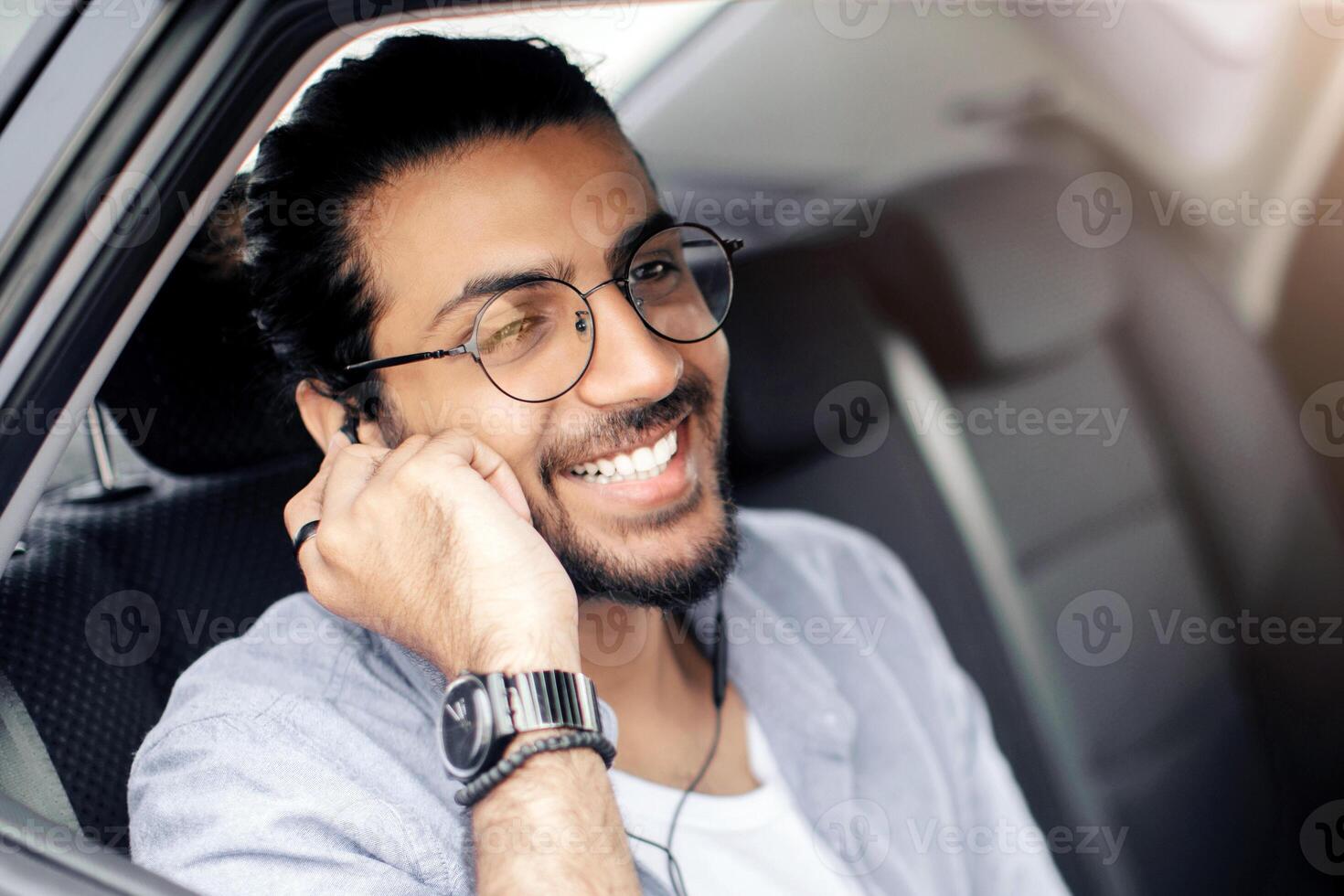 Cheerful arab guy sitting at car back seat, listening to music while going somewhere by transportation service, using earphones and mobile application on smartphone, enjoying view, copy space photo