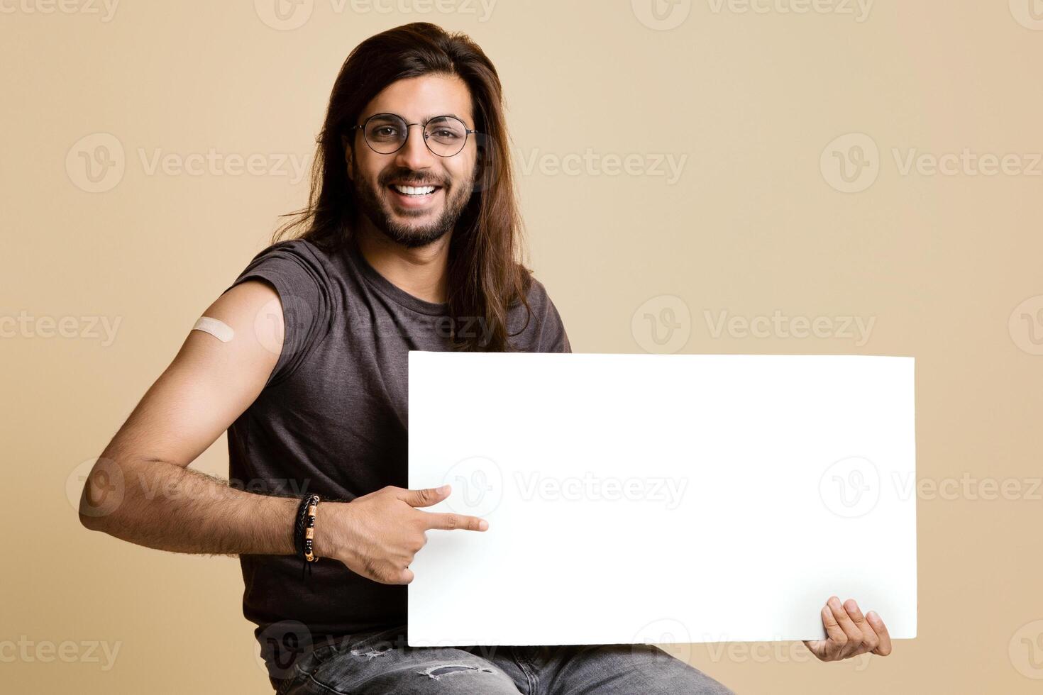Handsome middle-eastern young man with long hair and glasses got vaccinated against COVID-19, holding horizontal white blank board, sitting on chair over beige studio background, copy space, mockup photo