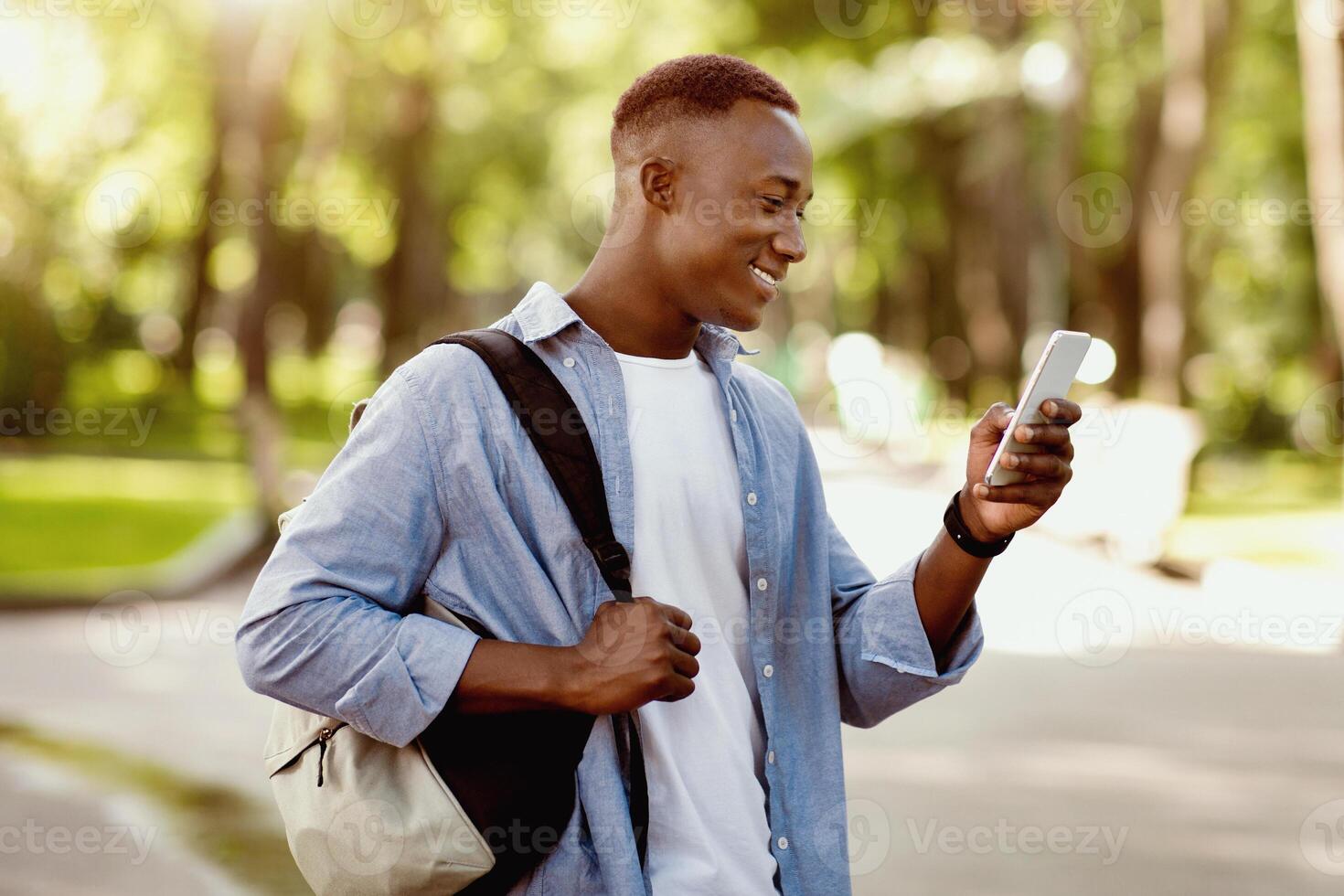 Cheerful black man with backpack using cellphone on walk in city park. African American man checking mail, sending message, watching entertainment , browsing internet on mobile phone outdoors photo