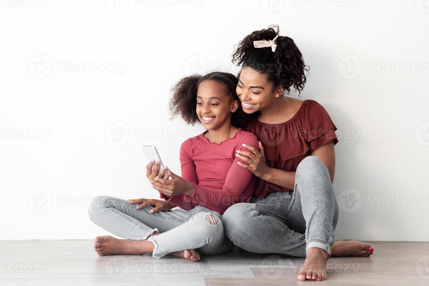 Beautiful black mother and daughter using mobile phone together, sitting on floor, hugging and taking selfie for social media, enjoying time together at home, white empty wall backgound, copy space photo