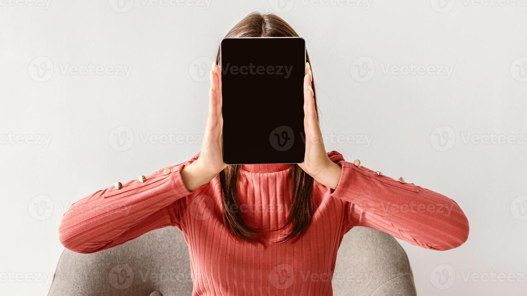 Young woman holding tablet pc with blank screen in front of her face on white studio background, mockup for app or website. Millennial lady presenting space for advertisement on touch pad photo