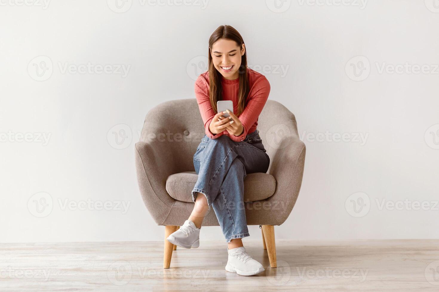 Happy Caucasian woman using cellphone, chatting on internet, working or studying online, sitting in armchair against white studio wall, full length. Cheery young lady watching on mobile phone photo