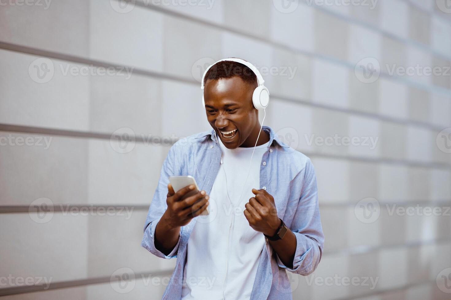 Victorious black man with headphones making YES gesture while looking at his mobile phone near brick wall photo