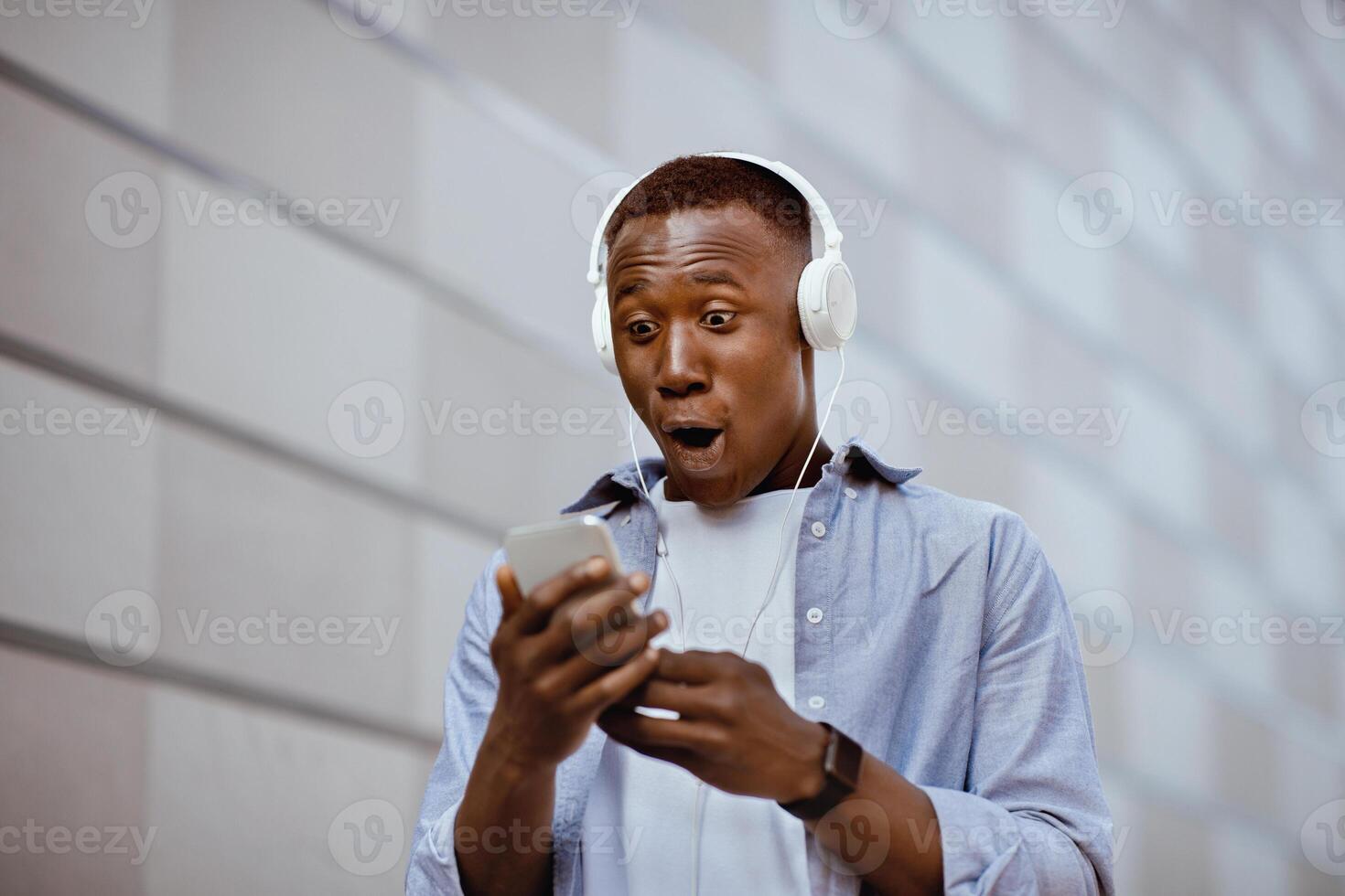 Shocked black guy in headphones looking at cellphone screen near brick wall on city street photo