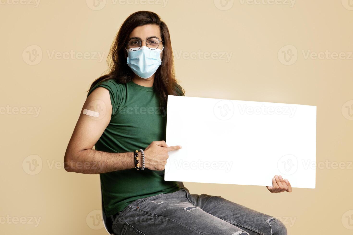 Middle-eastern vaccinated man in protective face mask with empty board posing on beige studio background. Long-haired young arab guy recommending vaccination while COVID-19 pandemic photo