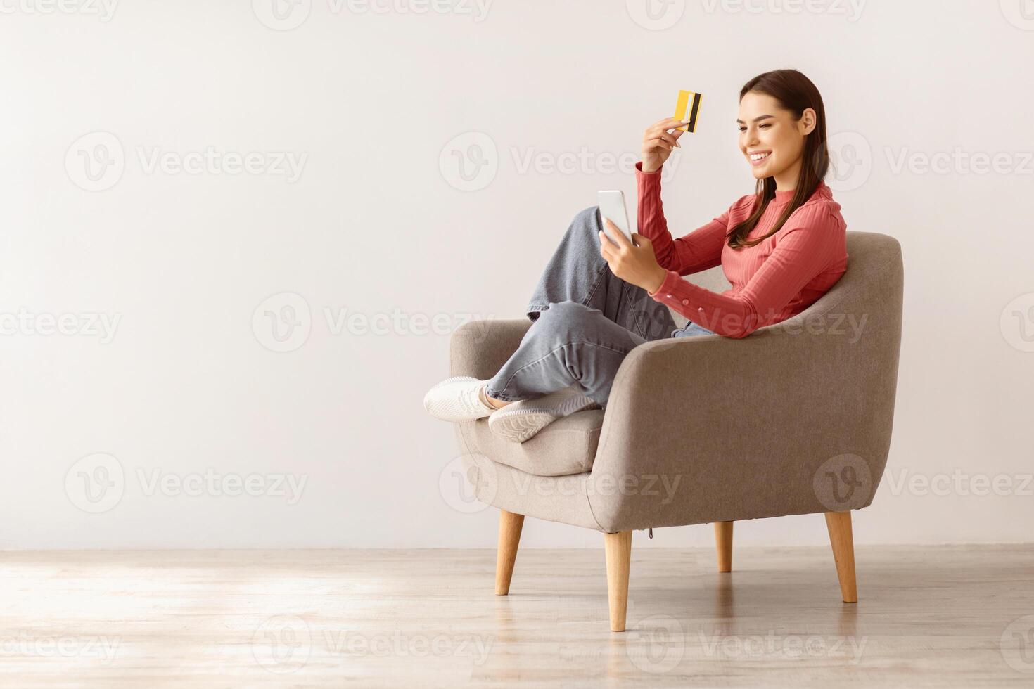 Online shopping. Lovely Caucasian lady with smartphone and credit card sitting in armchair, buying goods in web store, copy space. Young woman purchasing things on internet photo