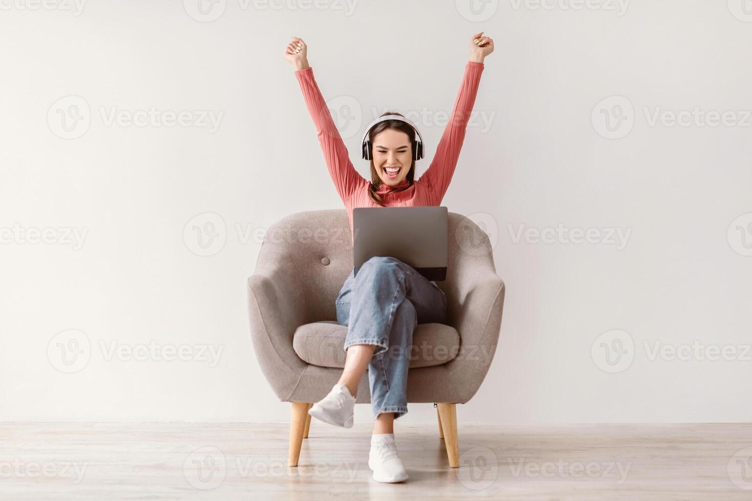 Full length of excited young lady with laptop and headphones raising her arms up, winning online casino bet, shopping on sale, reaching goal, achieving success, sitting in armchair against white wall photo