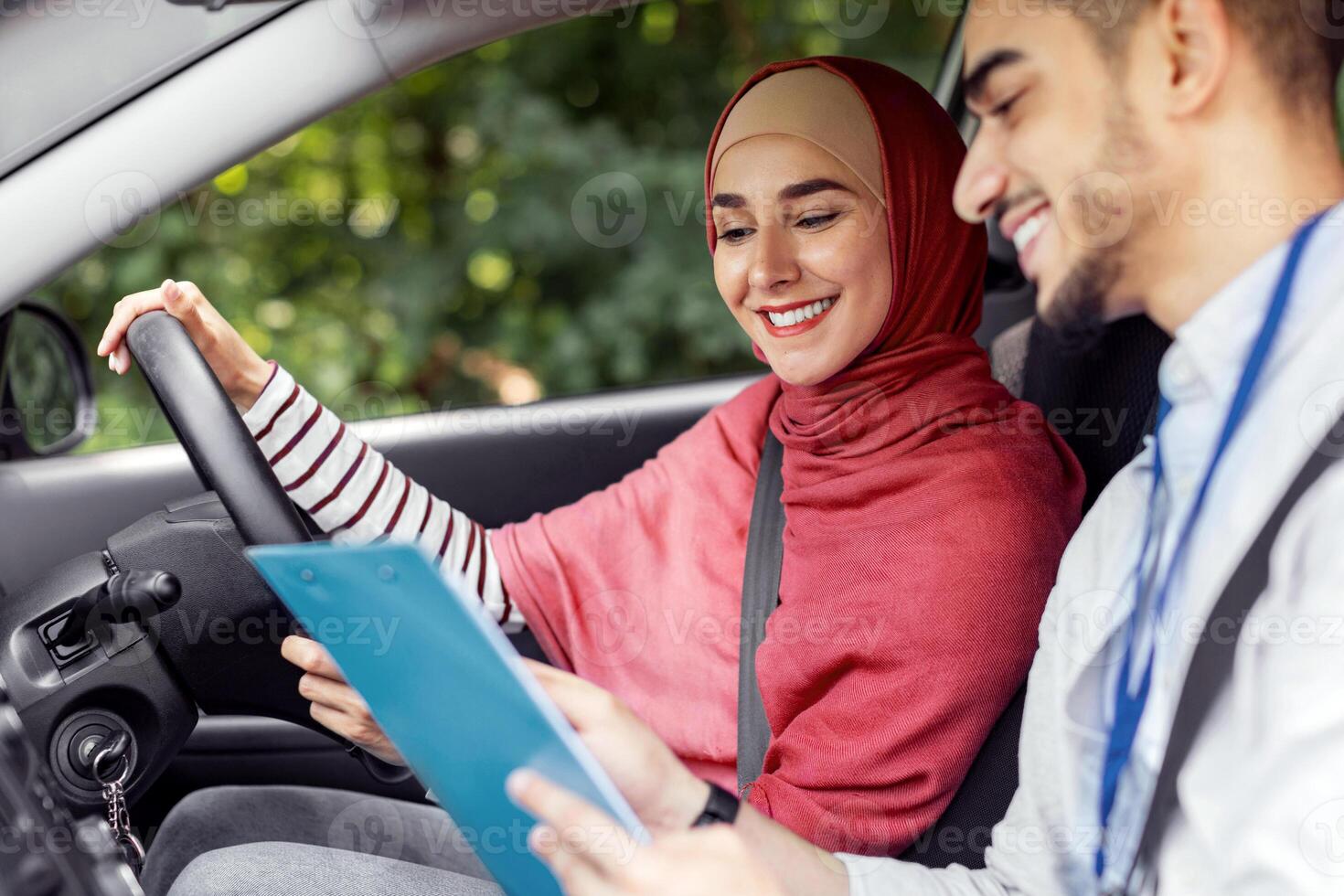 Smiling millennial middle eastern man, explain rules, put mark for driving test for woman in hijab at steering wheel at car, outdoor. Teacher teaches student to drive, driving class, exam in traffic photo