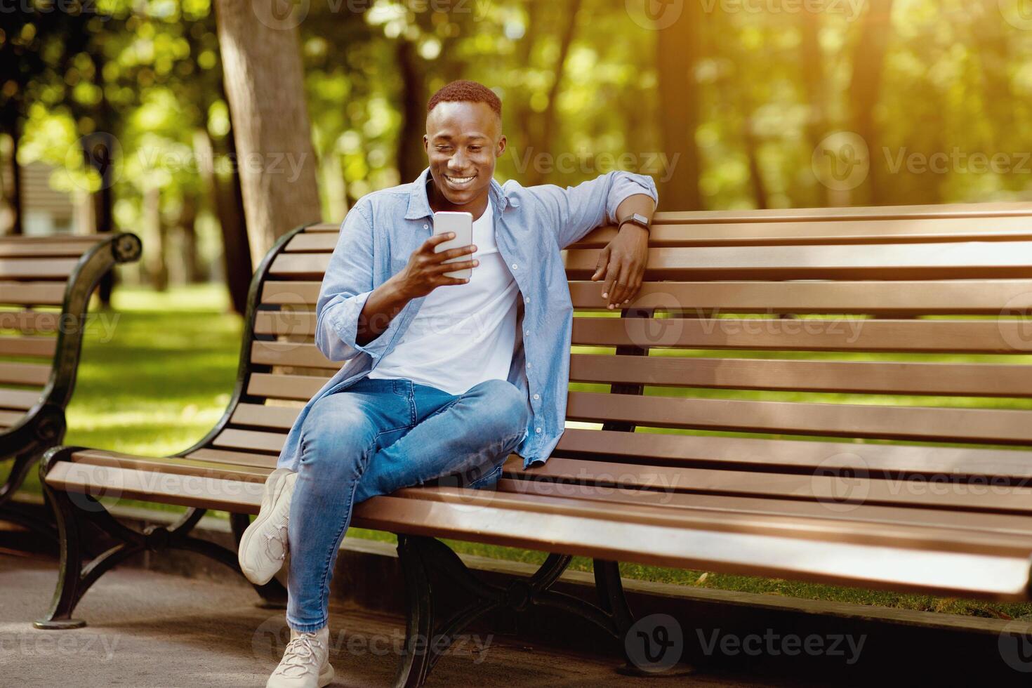 Smiling black man sitting on bench at park and using his smartphone, copy space photo