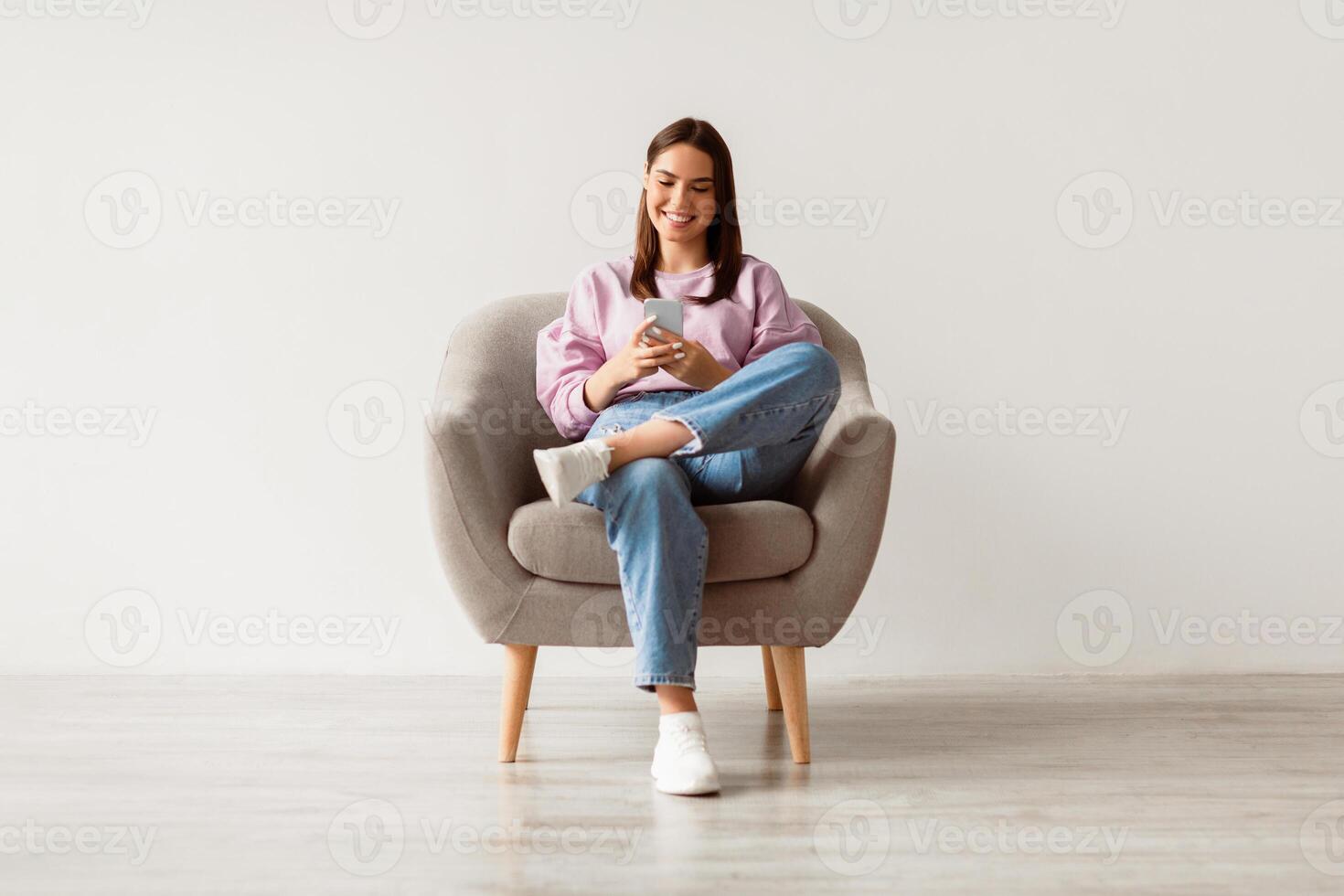 Beautiful young woman sitting in armchair, looking at mobile phone screen, using new cool application against white studio wall, full length. Joyful lady using smartphone for work or entertainment photo