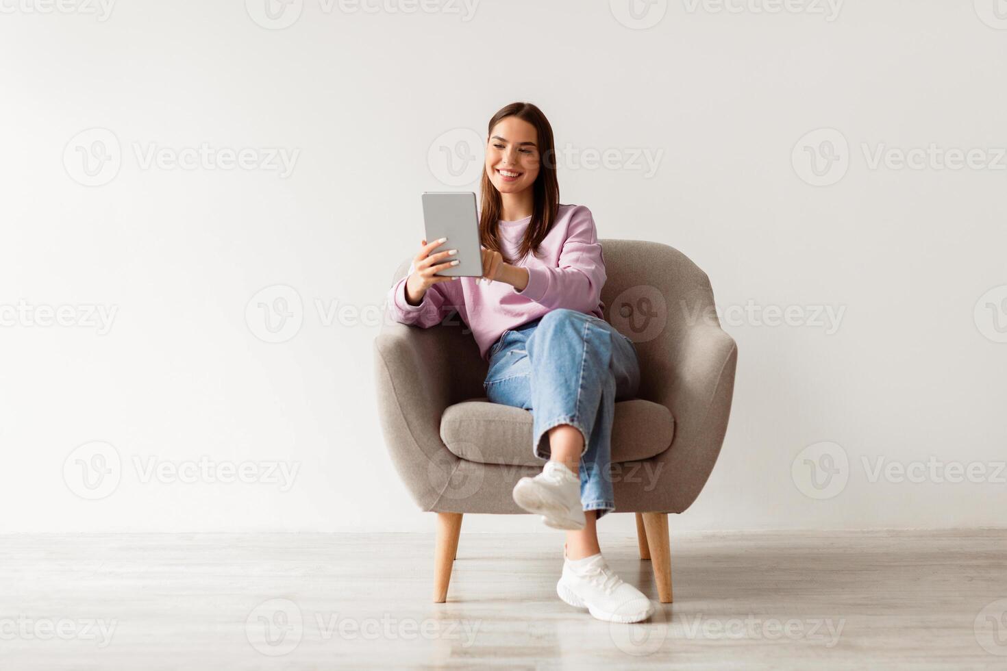Modern technologies and communications. Full length of cheery young woman with tablet pc sitting in armchair, working or studying online, having chat against white studio wall photo